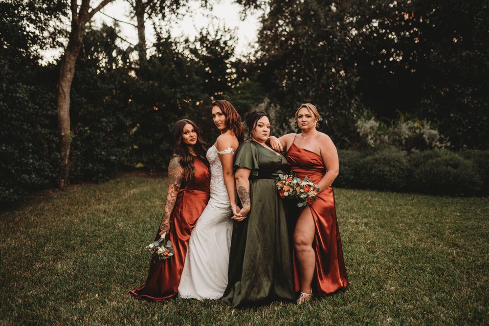 Bridesmaids in rust-colored dresses pose with bride on grassy lawn.