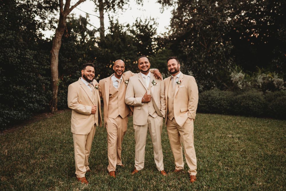 Groomsmen in beige suits stand together on grass during sunset wedding photos.