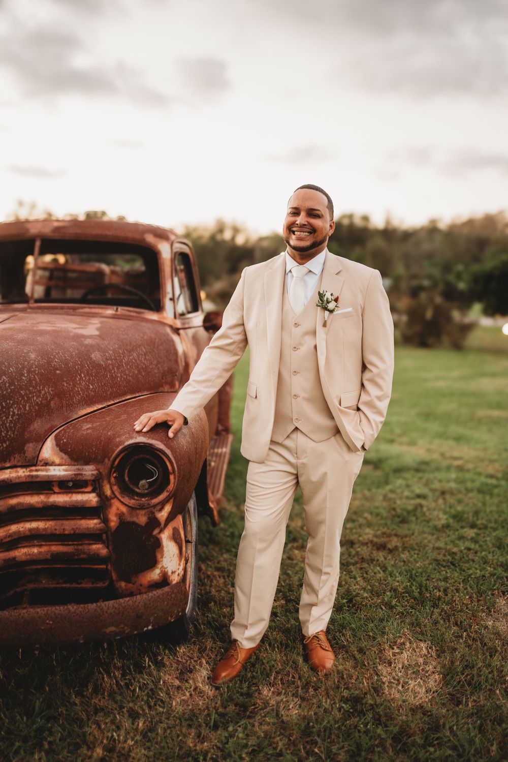 Groom in cream suit poses beside vintage rustic truck during wedding photoshoot.