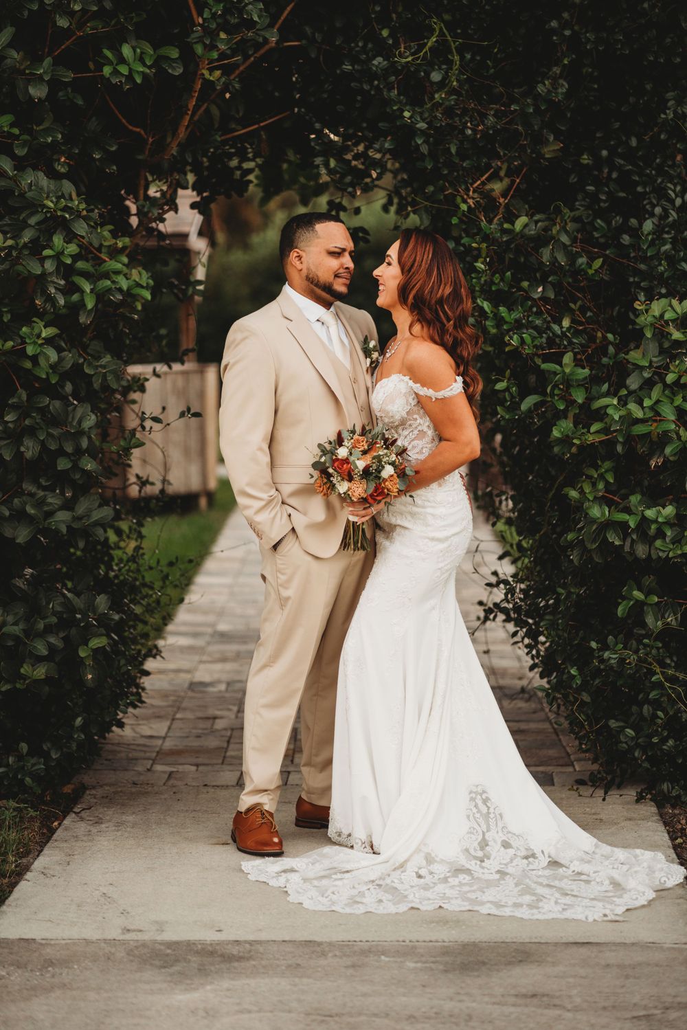 Elegant bride and groom pose together on garden pathway surrounded by lush greenery.