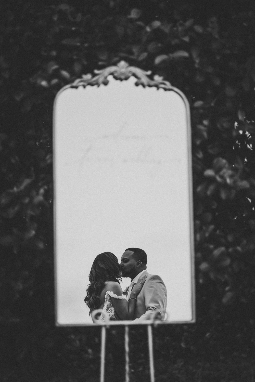 Artistic black and white wedding photo captured through decorative mirror frame outdoors.