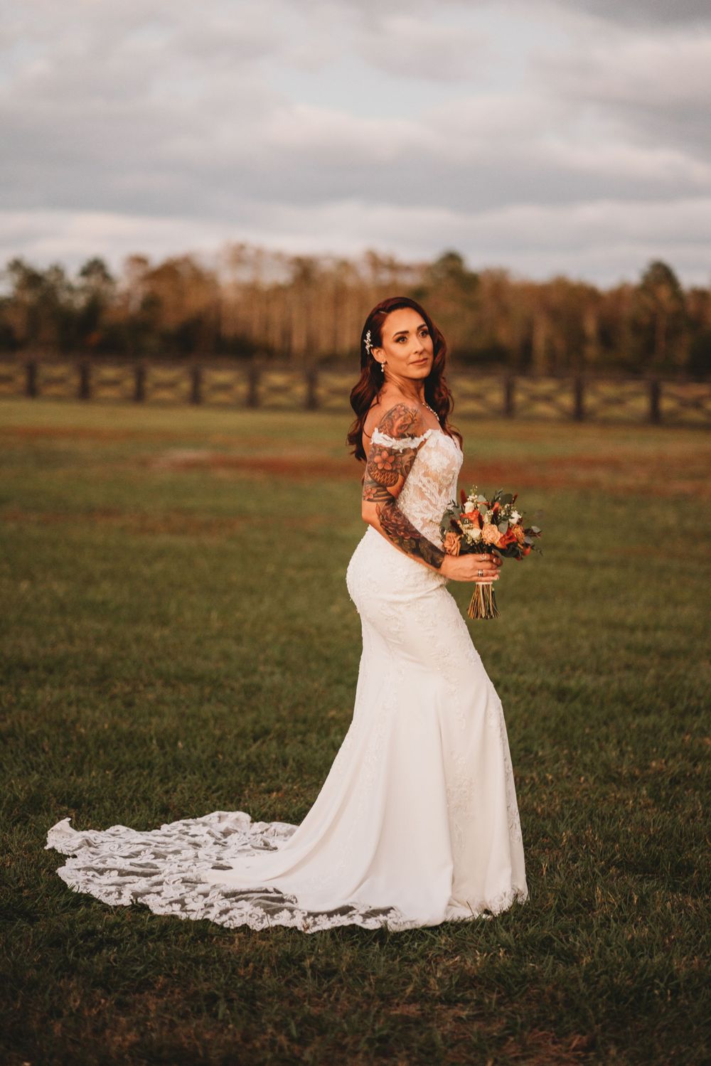 Bride in fitted lace gown with long train poses in rustic outdoor setting.