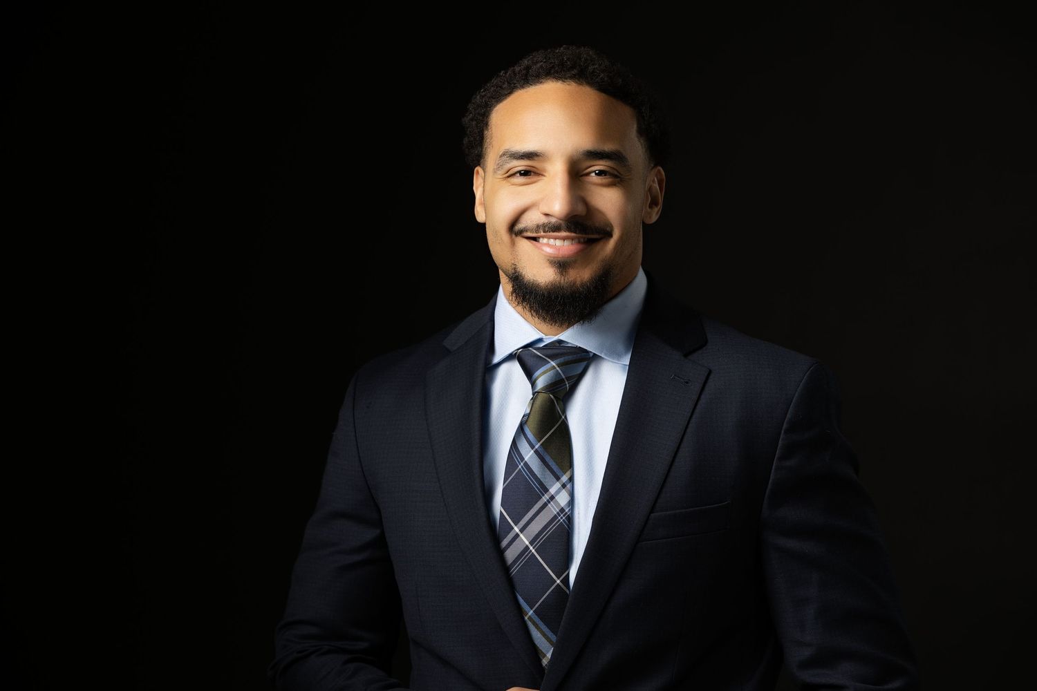 Professional headshot of person in dark suit against black background.