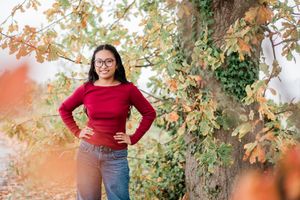 Senior portrait in a red sweater and jeans standing against an autumn tree with changing leaves in the background.