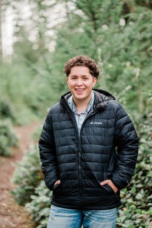 Outdoor senior portrait of boy wearing a black Columbia puffer jacket and jeans standing on a wooded trail.