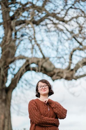 Person in a brown cable knit sweater stands peacefully under a large bare tree against a bright sky.