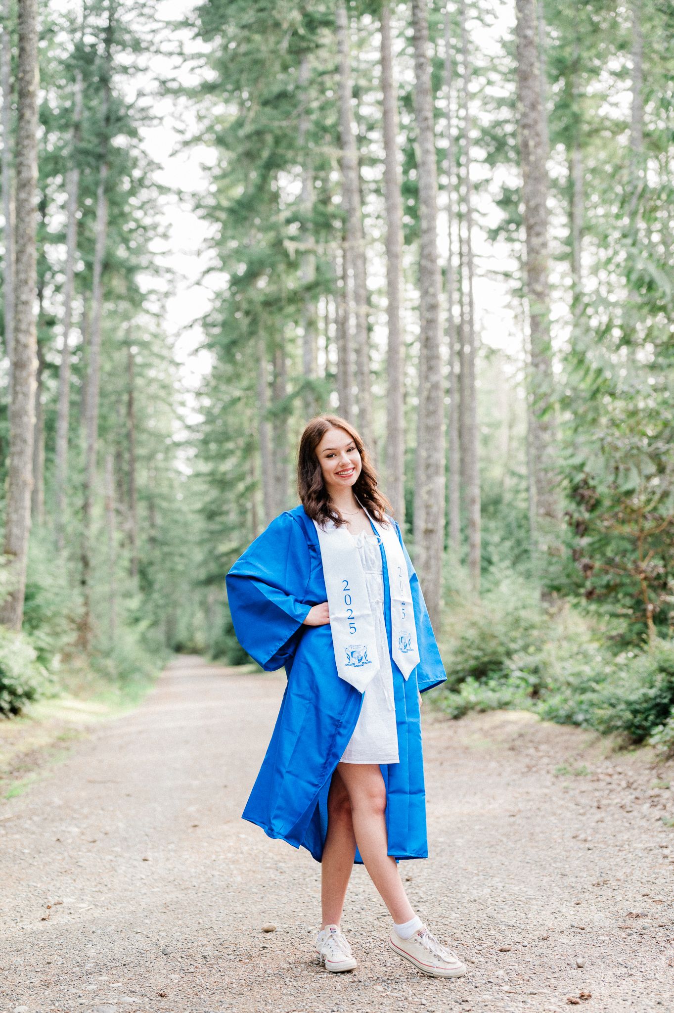 An Olympic High School Senior poses playfully on a forest path surrounded by tall evergreen trees.