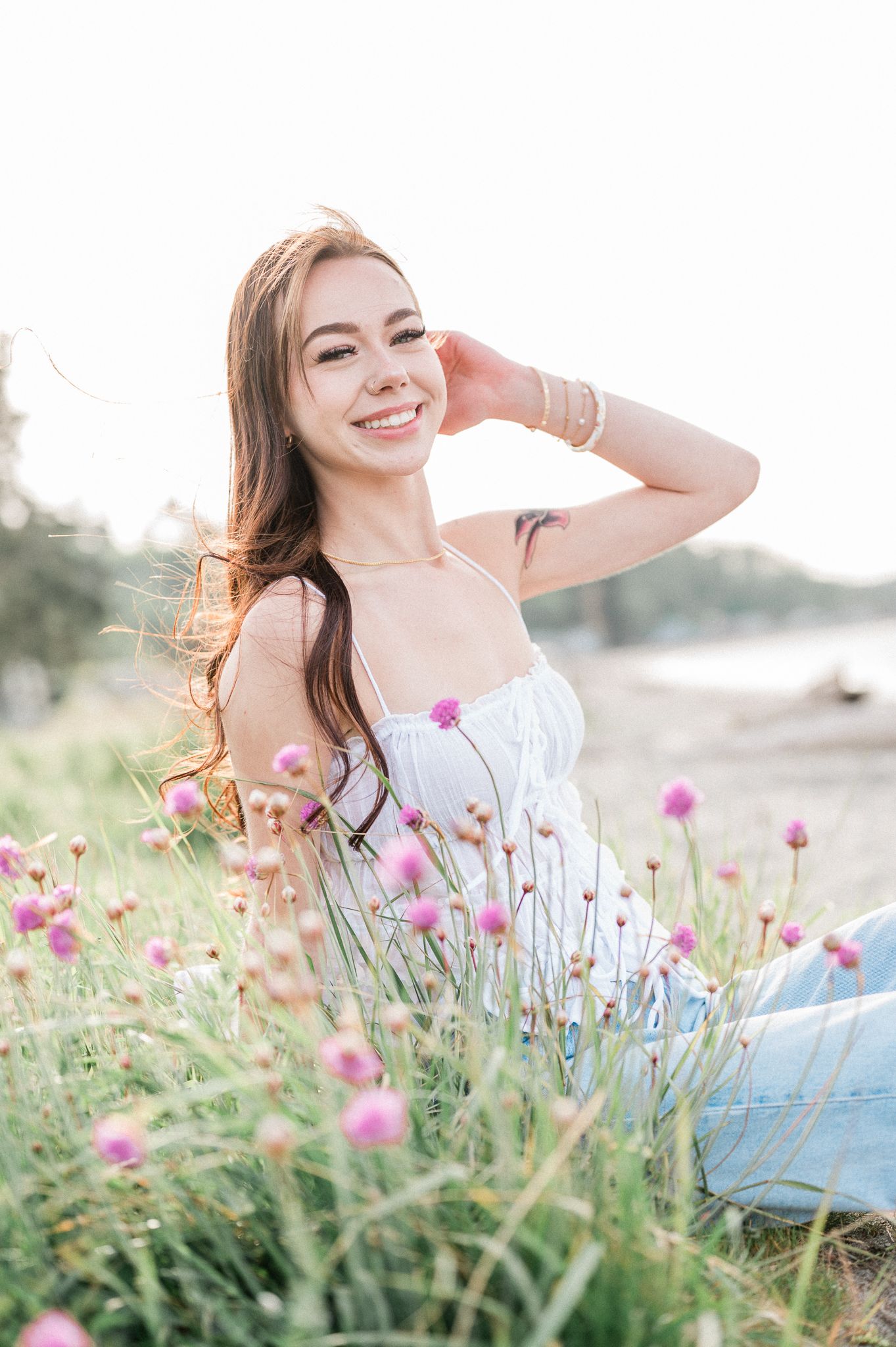 A high school senior in a white top sits gracefully among blooming pink wildflowers in a dreamy outdoor setting.