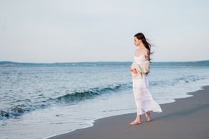 A senior portrait of a girl in a flowing white dress walking along a sandy beach with ocean waves and blue sky in the background.