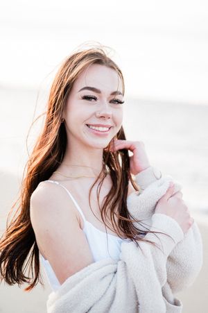 Girl in a white sweater and camisole poses against ocean at sunset for senior photos