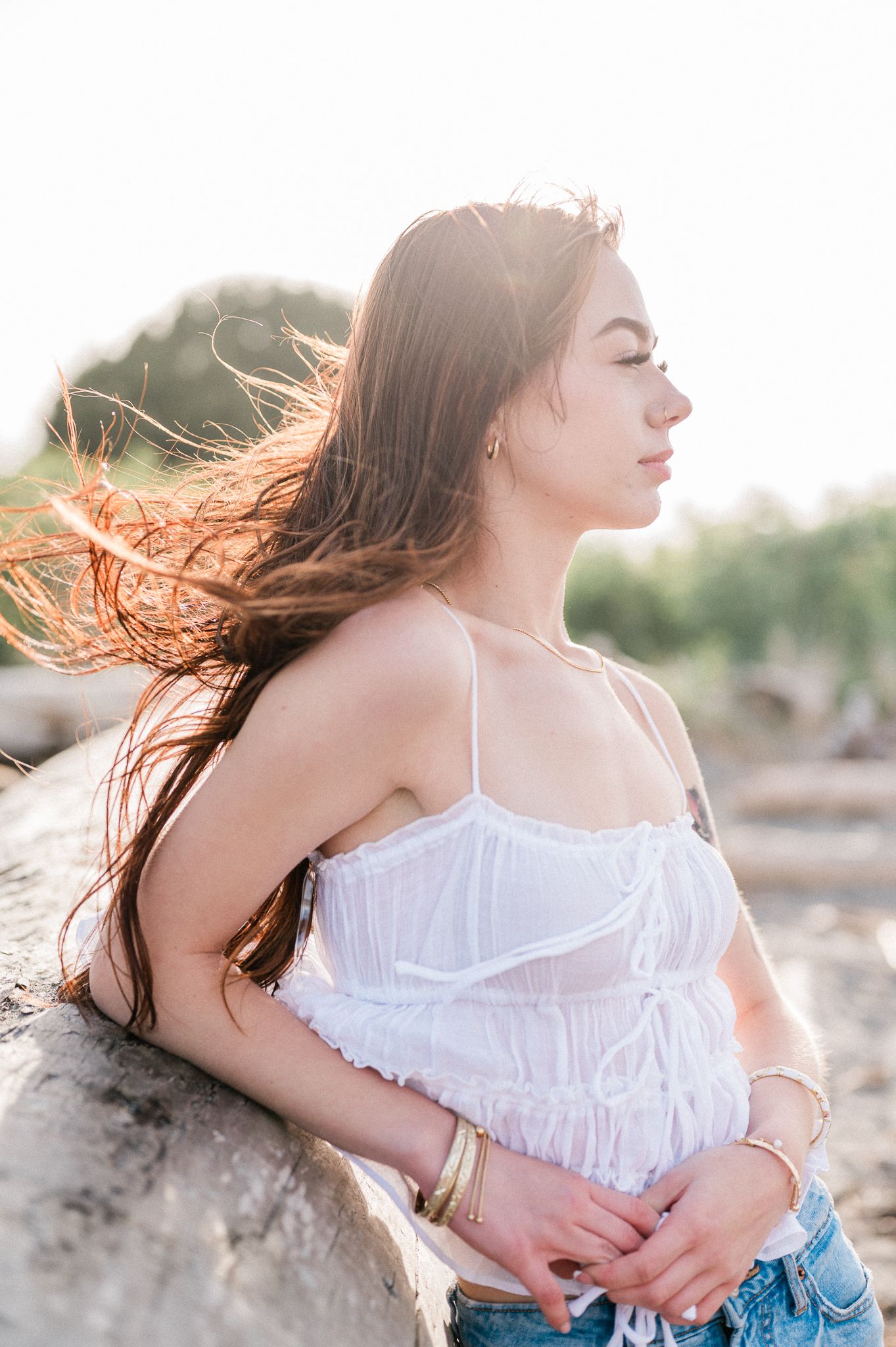 A girl in a white summer top and denim stands outdoors with windswept hair during golden hour lighting.