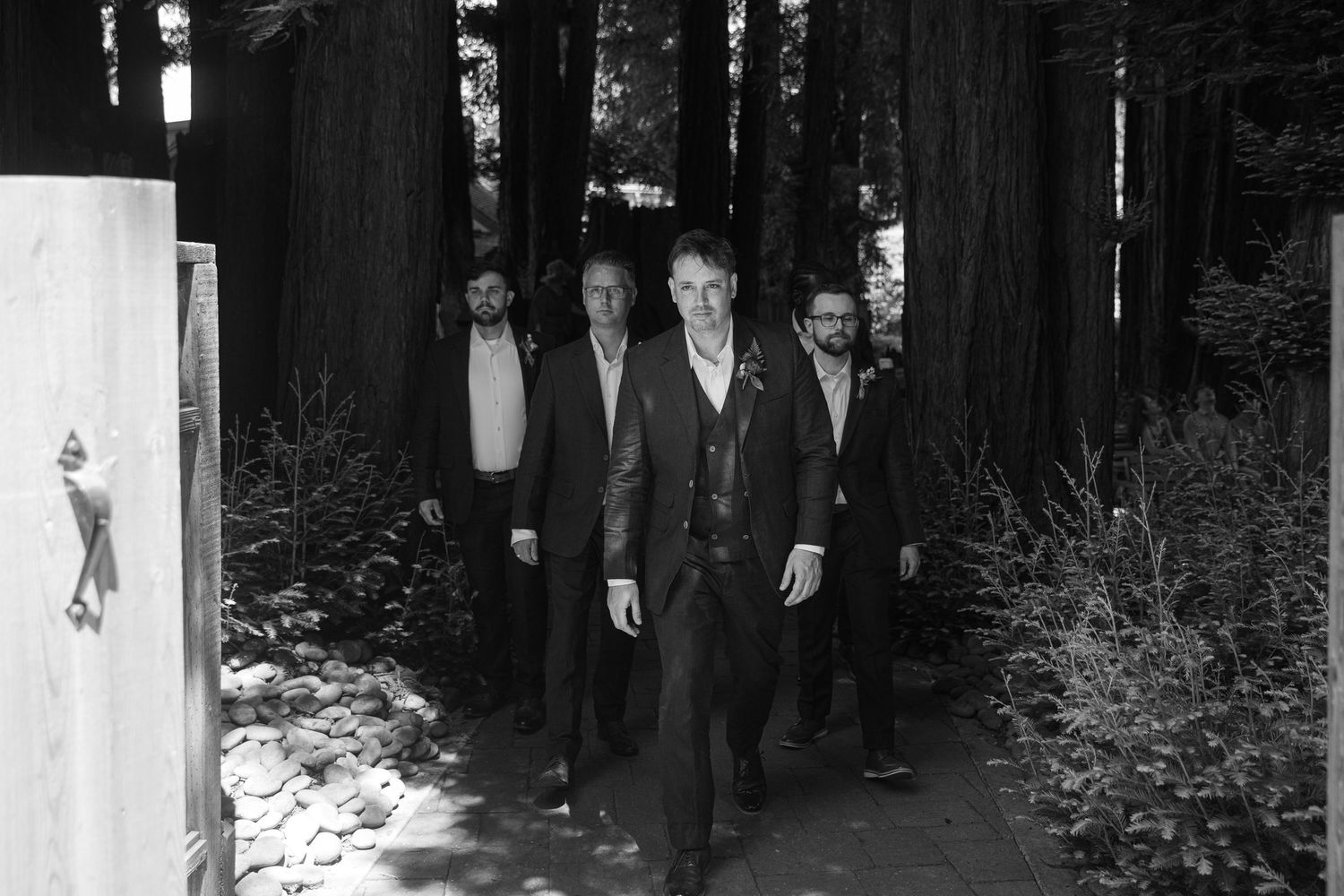 A black and white photograph shows a group of people walking through a dark wooded area.