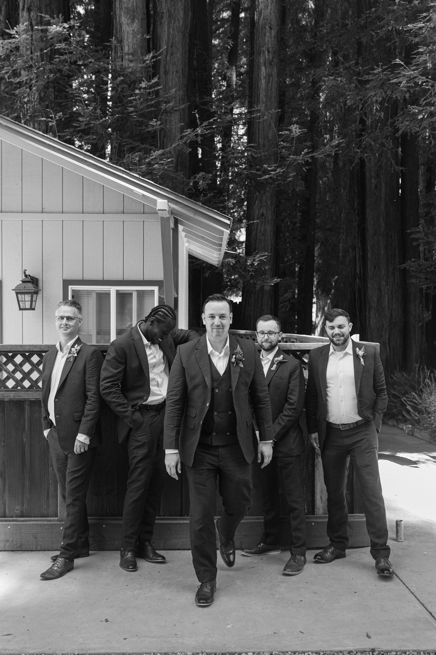 Groomsmen in dark suits pose playfully along a wall in a redwood forest setting.