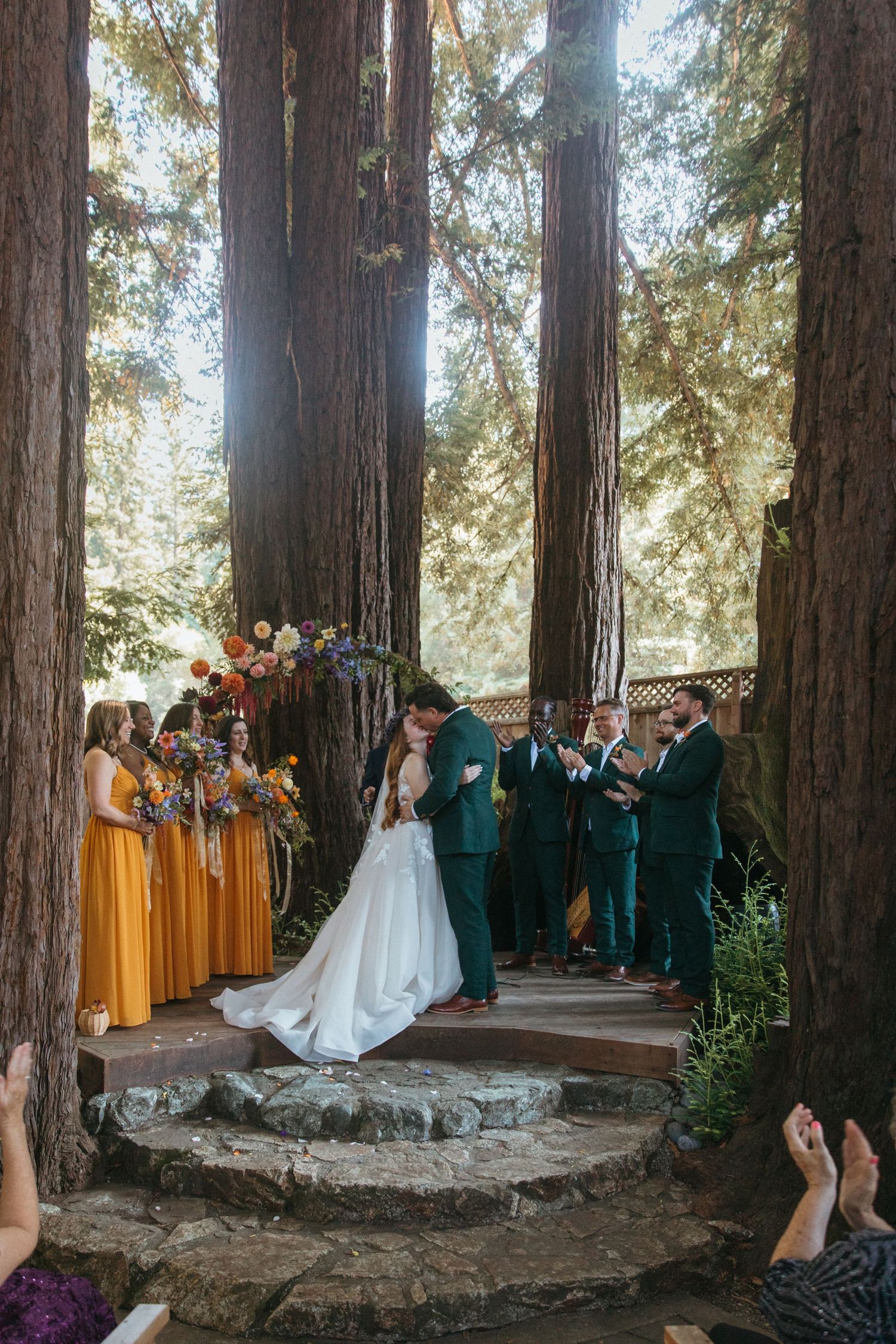A wedding ceremony takes place in a majestic redwood forest with bridesmaids in yellow dresses and groomsmen in green suits.
