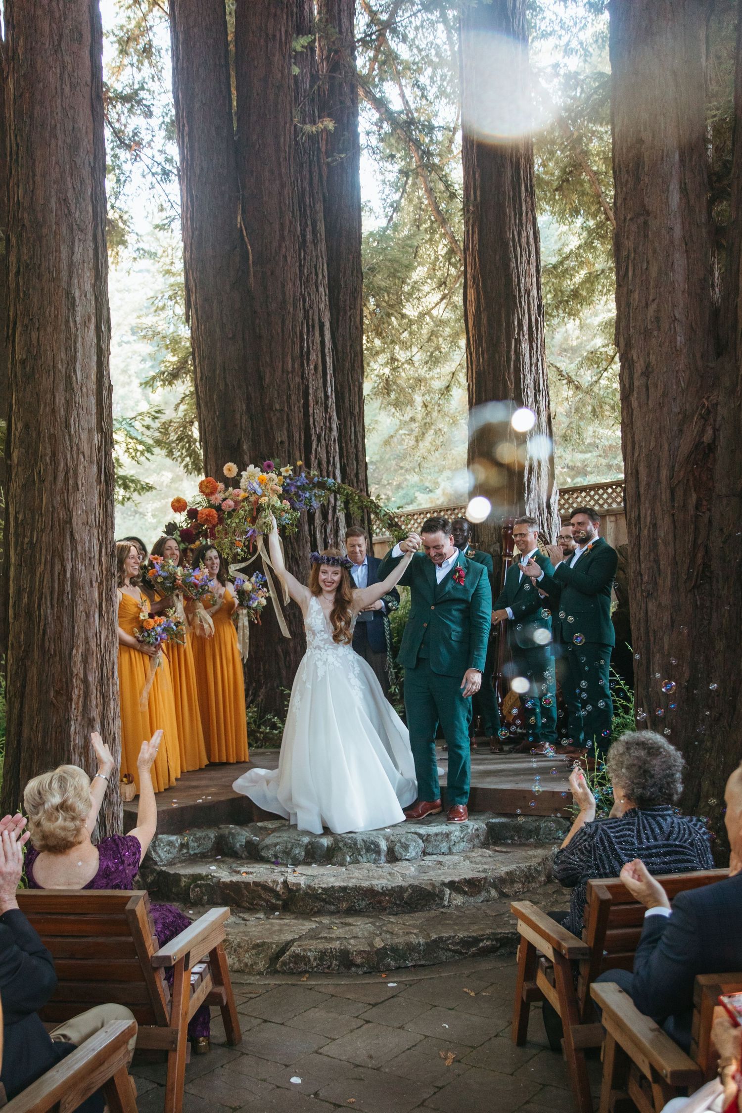 Wedding guests seated on wooden benches in a redwood grove during an outdoor ceremony.