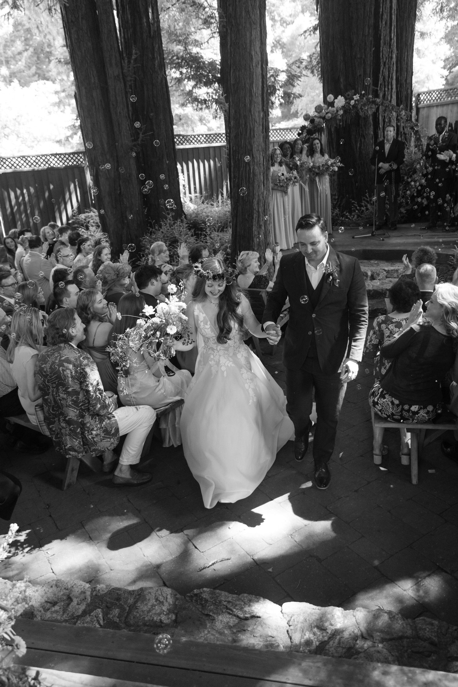 Wedding party dances and celebrates under tall redwood trees at an outdoor reception.