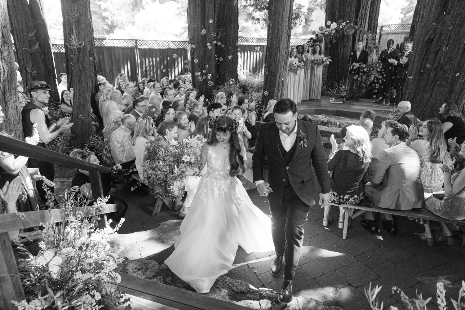 Black and white photo of wedding guests mingling at an outdoor forest reception with rustic decor.