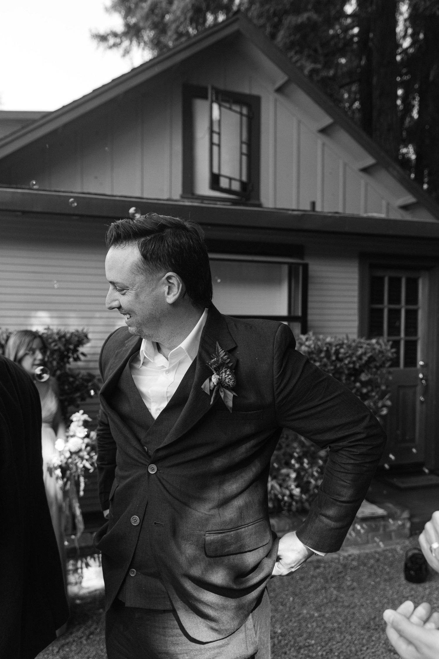 A black and white photo of someone in a suit jacket standing in front of a house with a floral boutonniere.