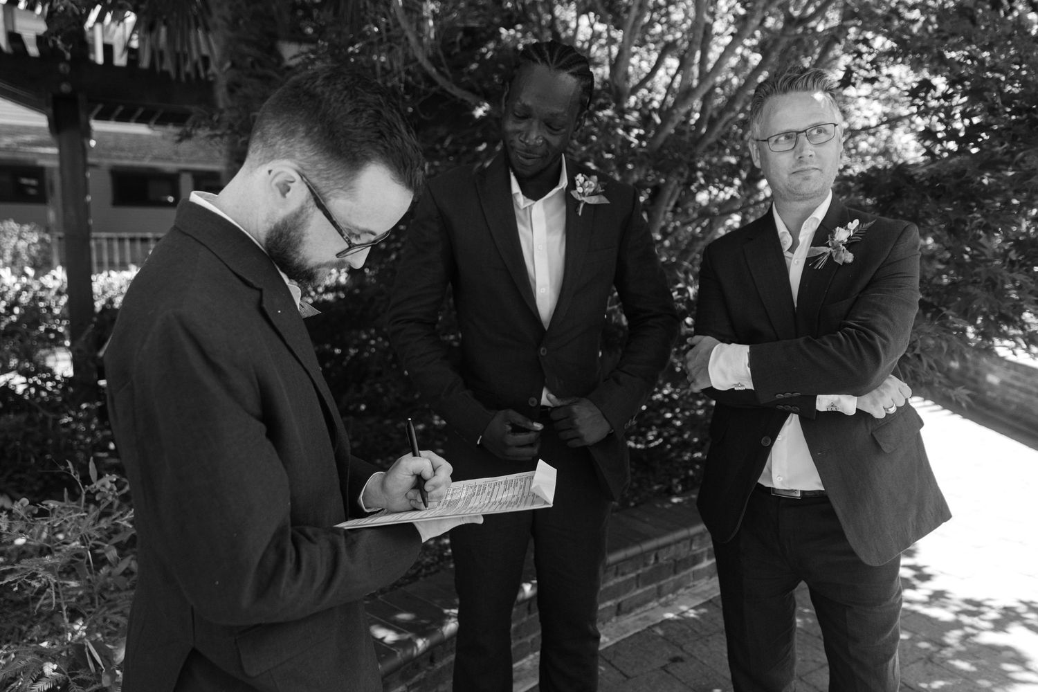 Wedding guests mingle during an outdoor celebration beneath tall evergreen trees in natural lighting.