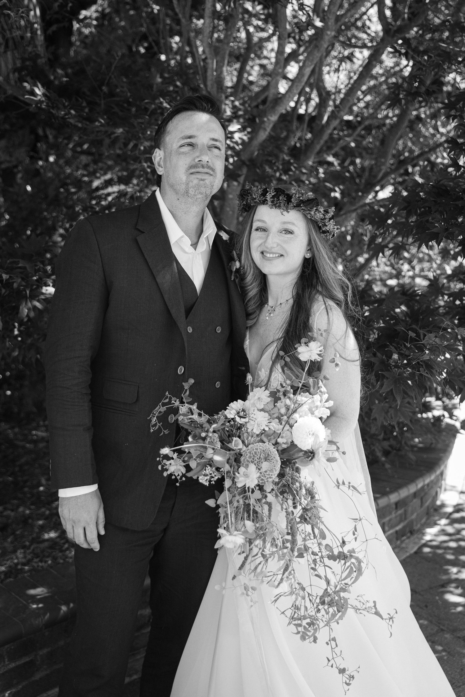 A black and white wedding photo shows a couple embracing outdoors under trees with the bride holding a floral bouquet.
