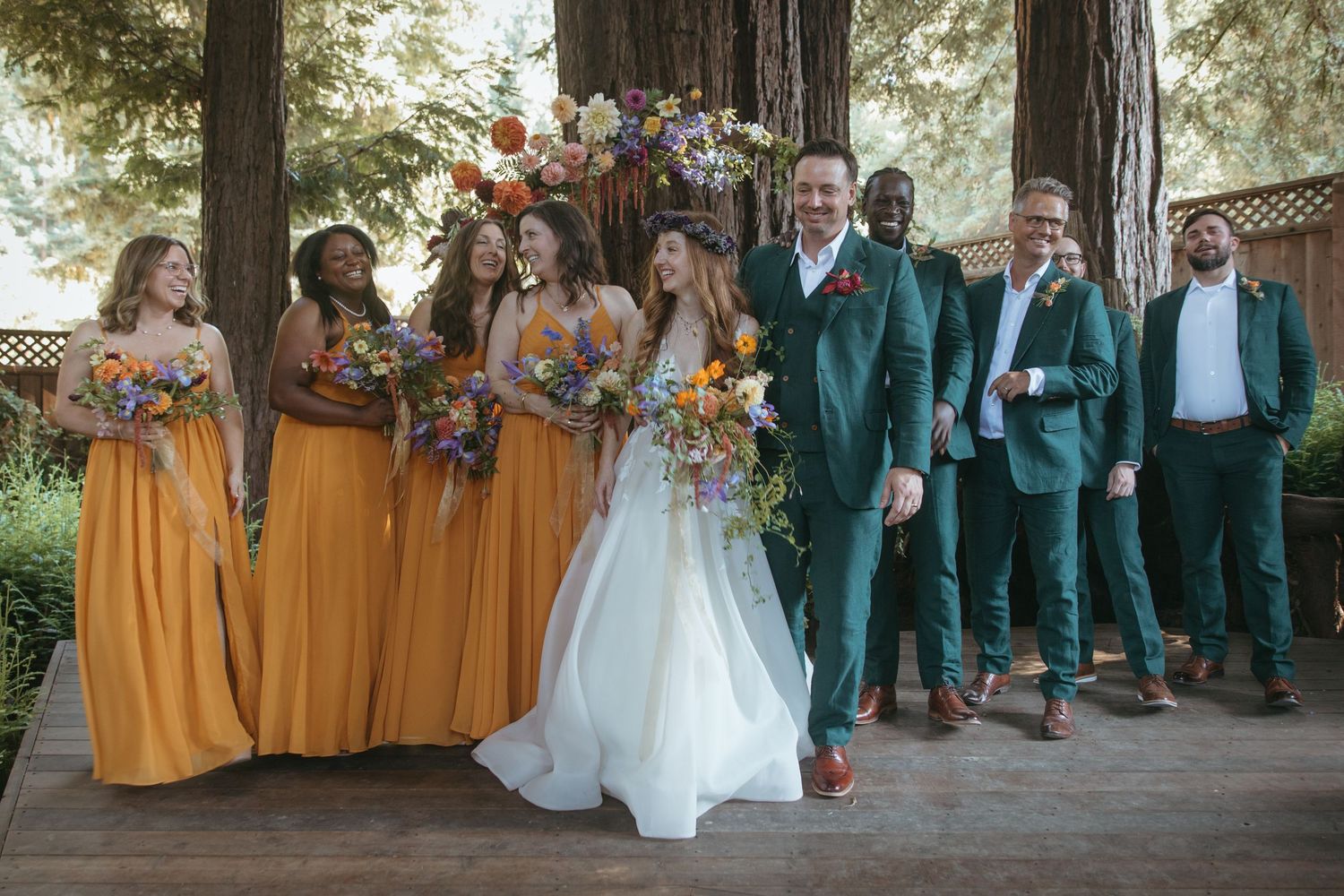 Wedding party members in teal suits and yellow dresses pose together in a forest setting for group photos.