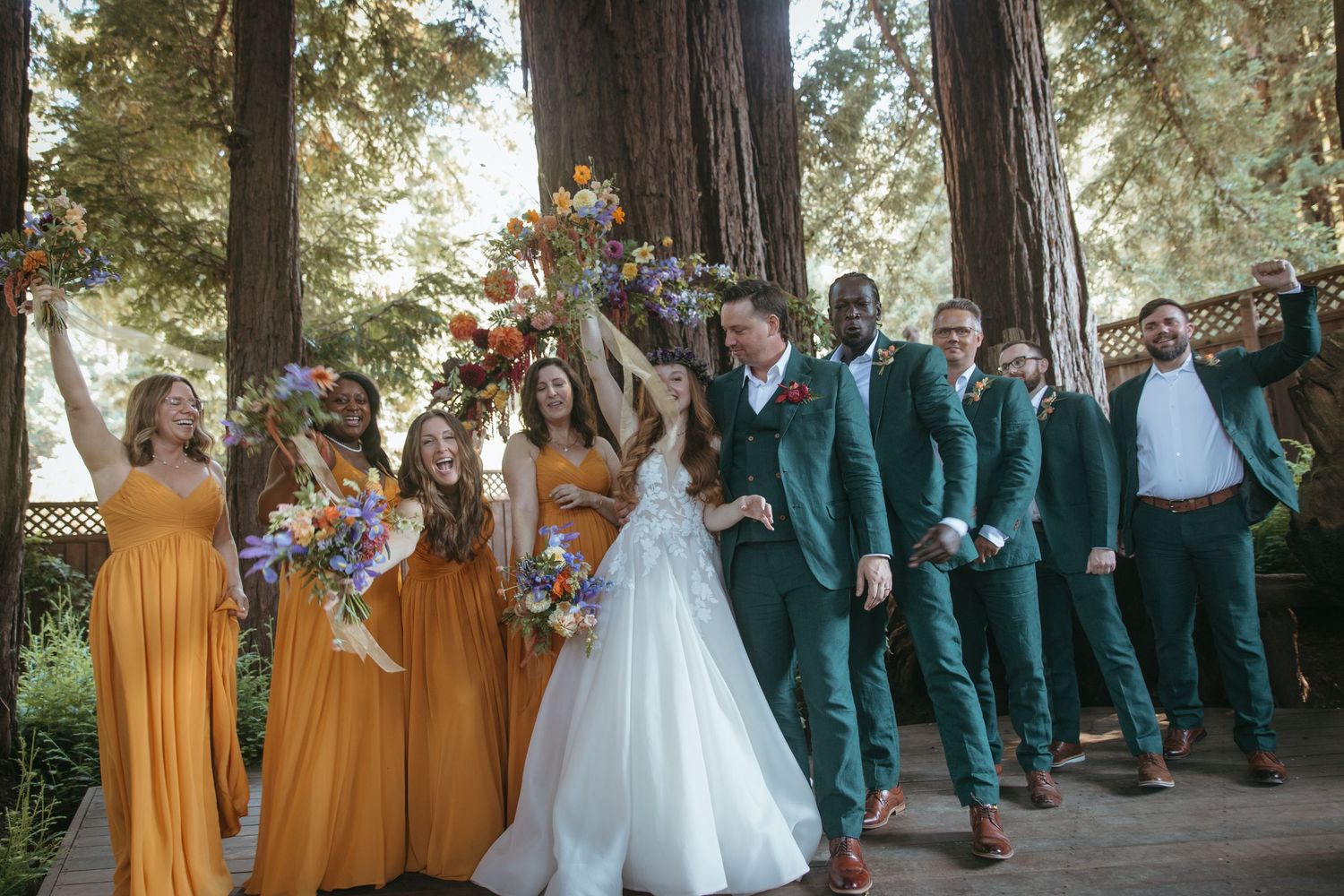 A joyful wedding celebration with attendants in emerald green and mustard yellow attire among towering redwoods.