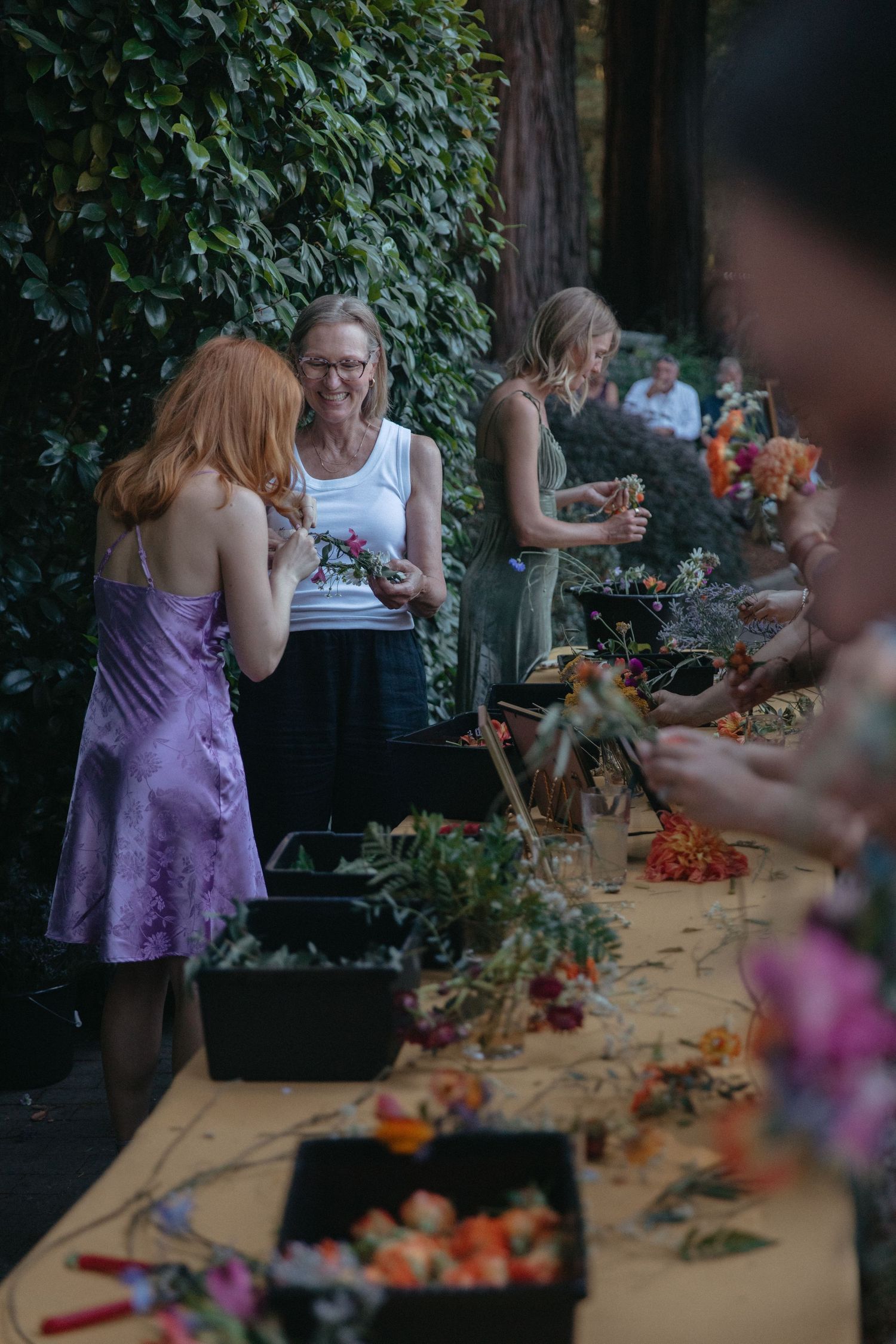 People working on floral arrangements at a rustic wooden table during a wedding preparation.