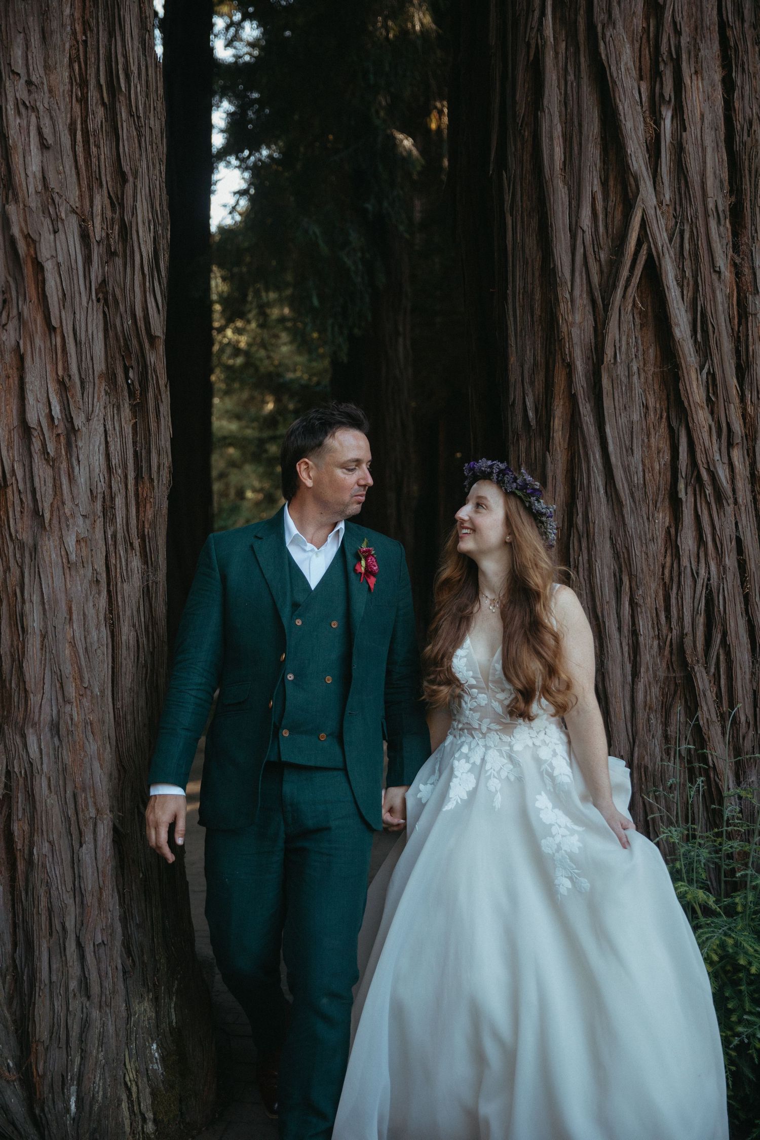 Wedding couple in green suit and white dress pose together among tall redwood trees.
