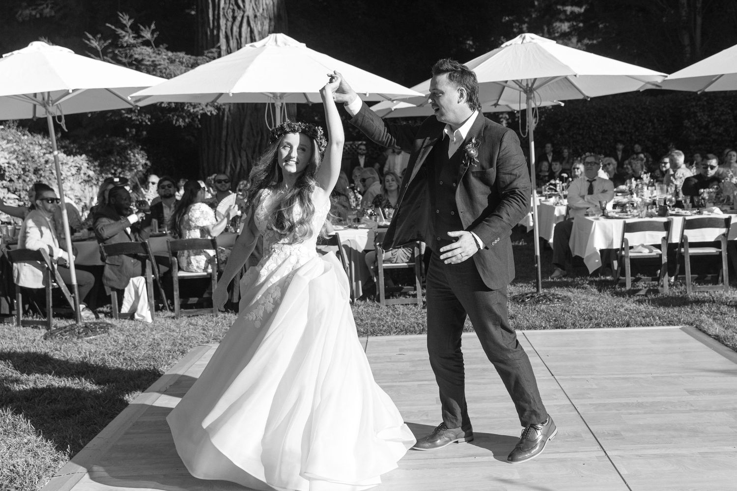 A black and white wedding photo captures a couple walking together on a paved path with umbrellas and trees in background.