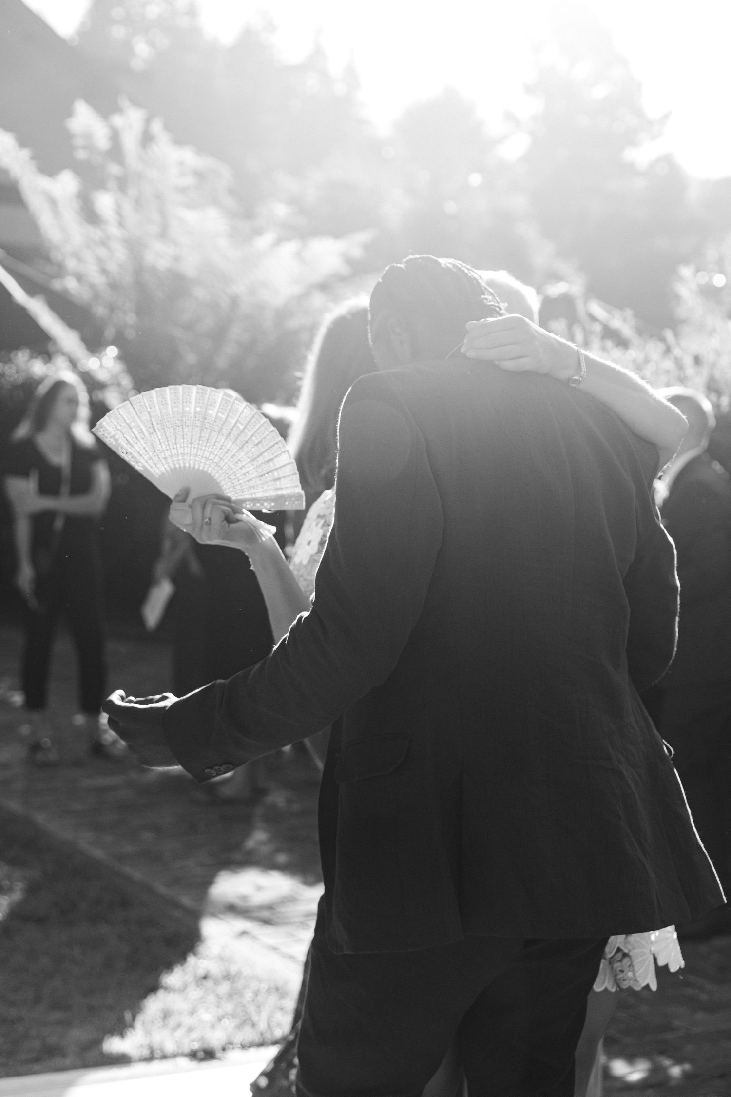 Silhouettes of wedding guests holding decorative fans while dancing outdoors in bright backlit sunlight.