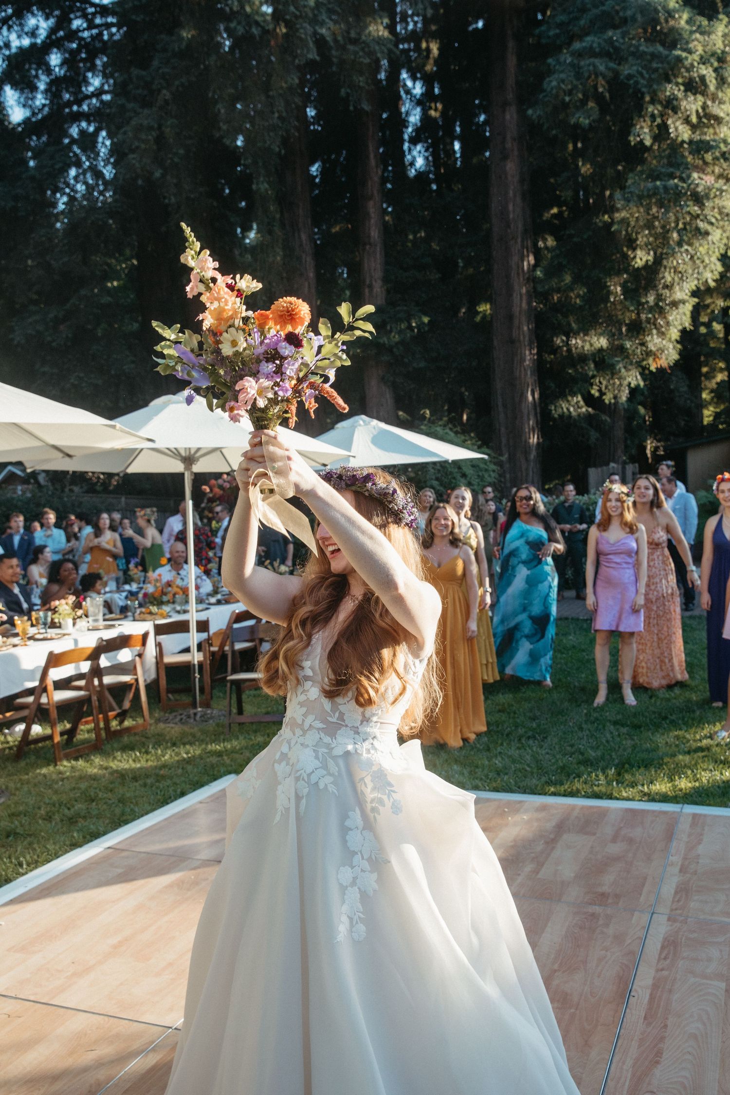 A bride in a white wedding gown tosses her bouquet during an outdoor forest wedding reception with guests watching.