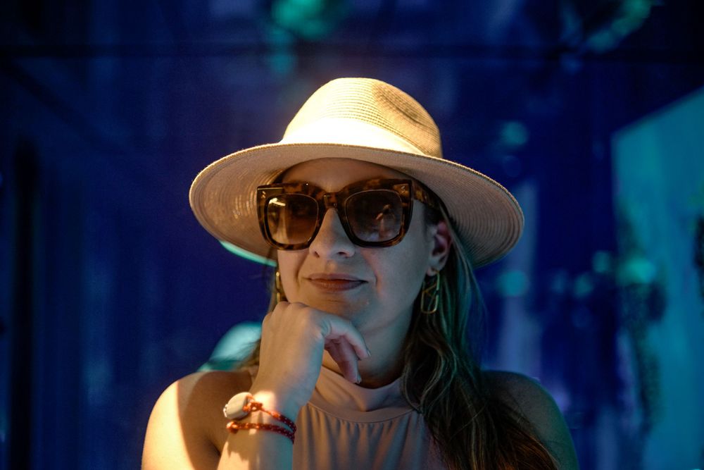 A person in a beige hat and sunglasses poses thoughtfully against a blue-tinted background.