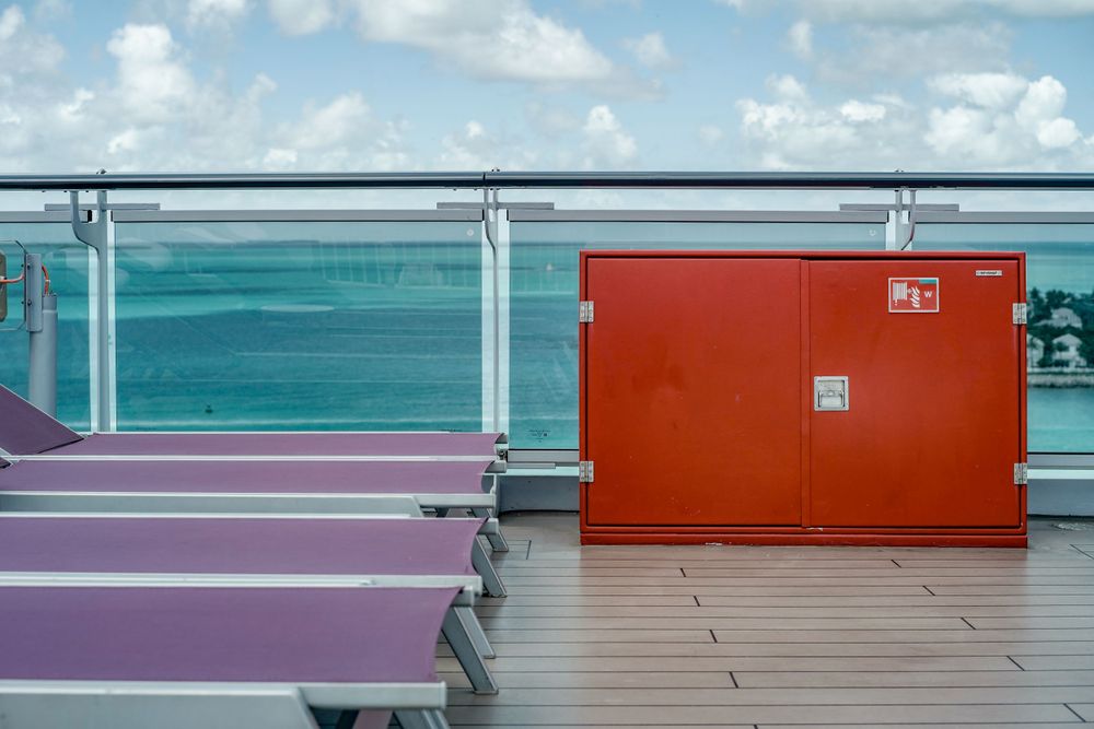 Cruise ship deck with purple loungers and a red service door against turquoise ocean waters.