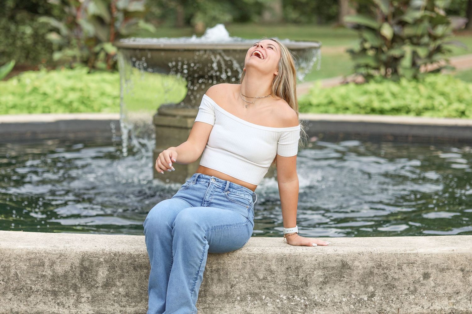 A casual summer outfit by a fountain features jeans and an off-shoulder white top.
