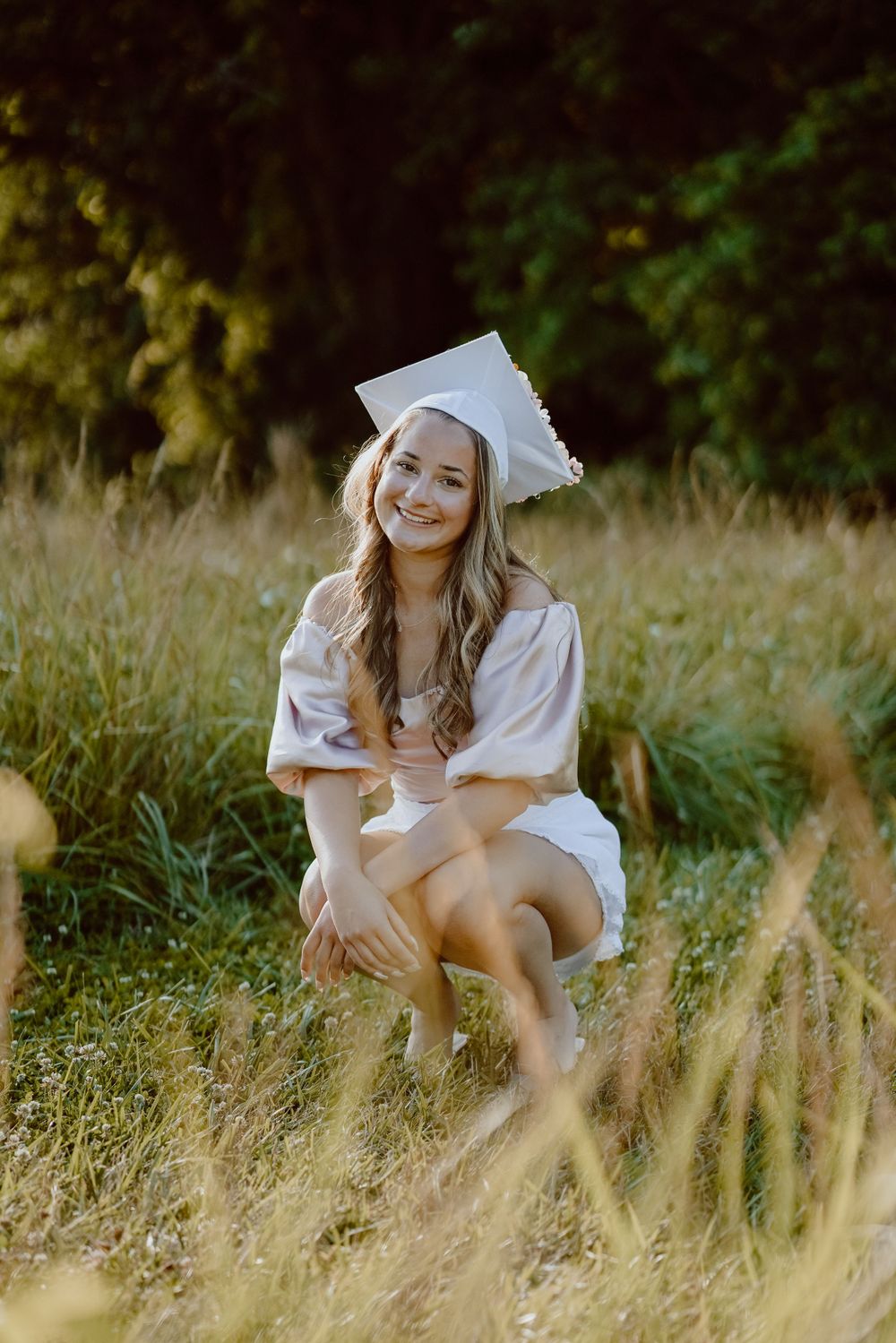 A graduate poses in white cap and gown while crouching in a sunlit meadow.