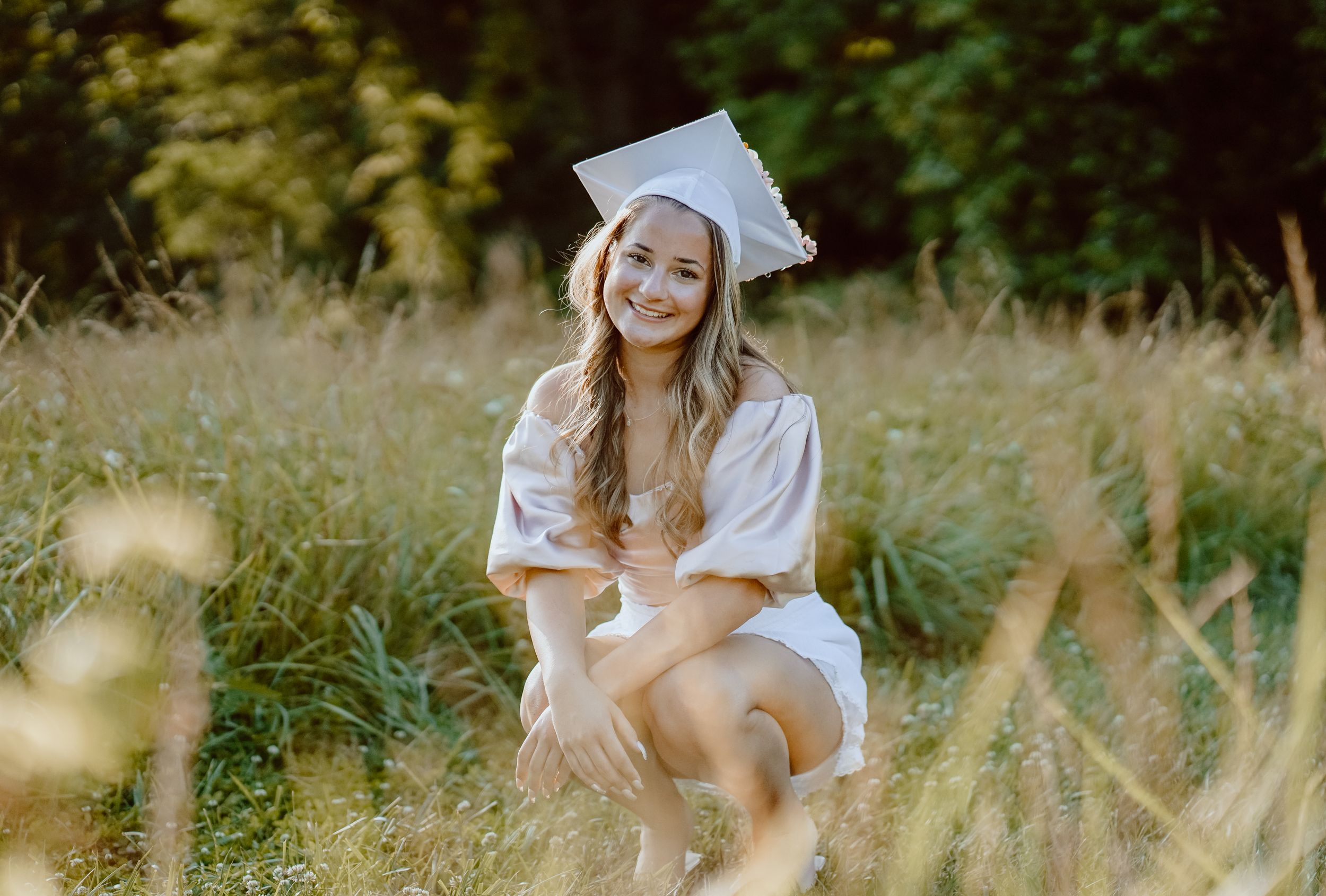 A graduation portrait in a white cap and gown sitting in tall summer grass.