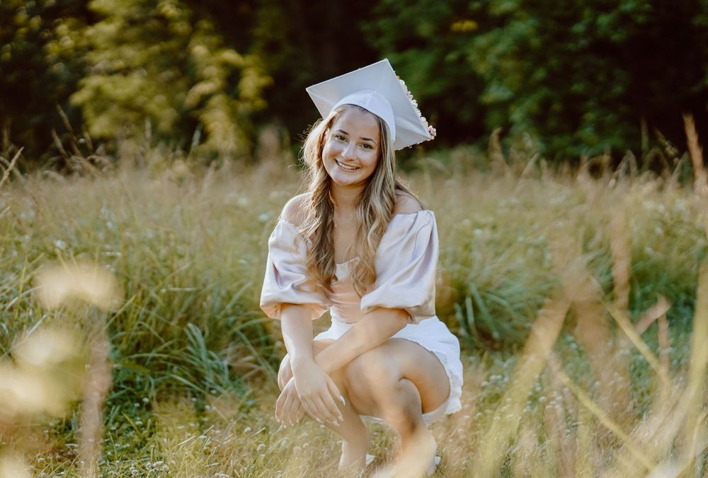 A graduation portrait in a white cap and gown sitting in tall summer grass.