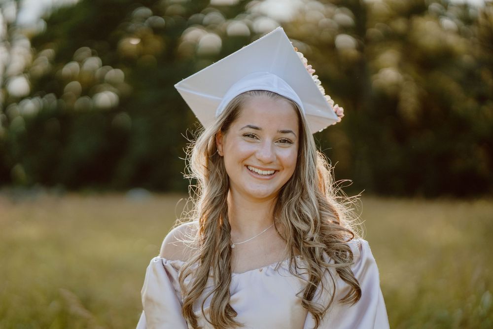 A graduate in white cap and gown smiles brightly in an outdoor portrait session.