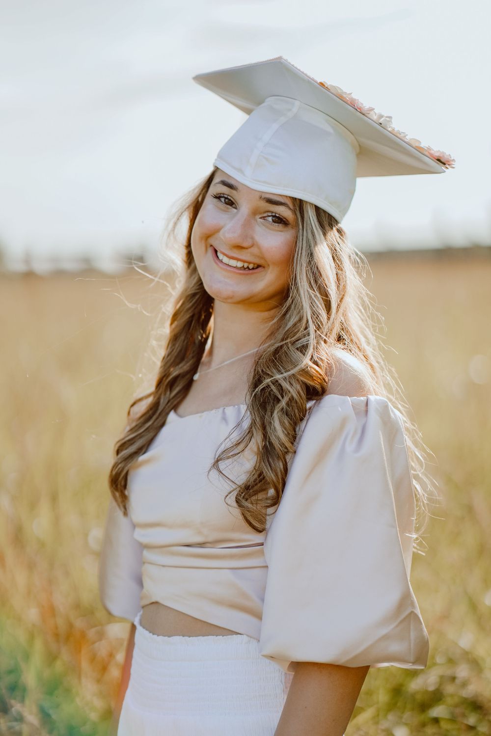 A graduate wears an off-shoulder white dress and cap while posing in a golden field.