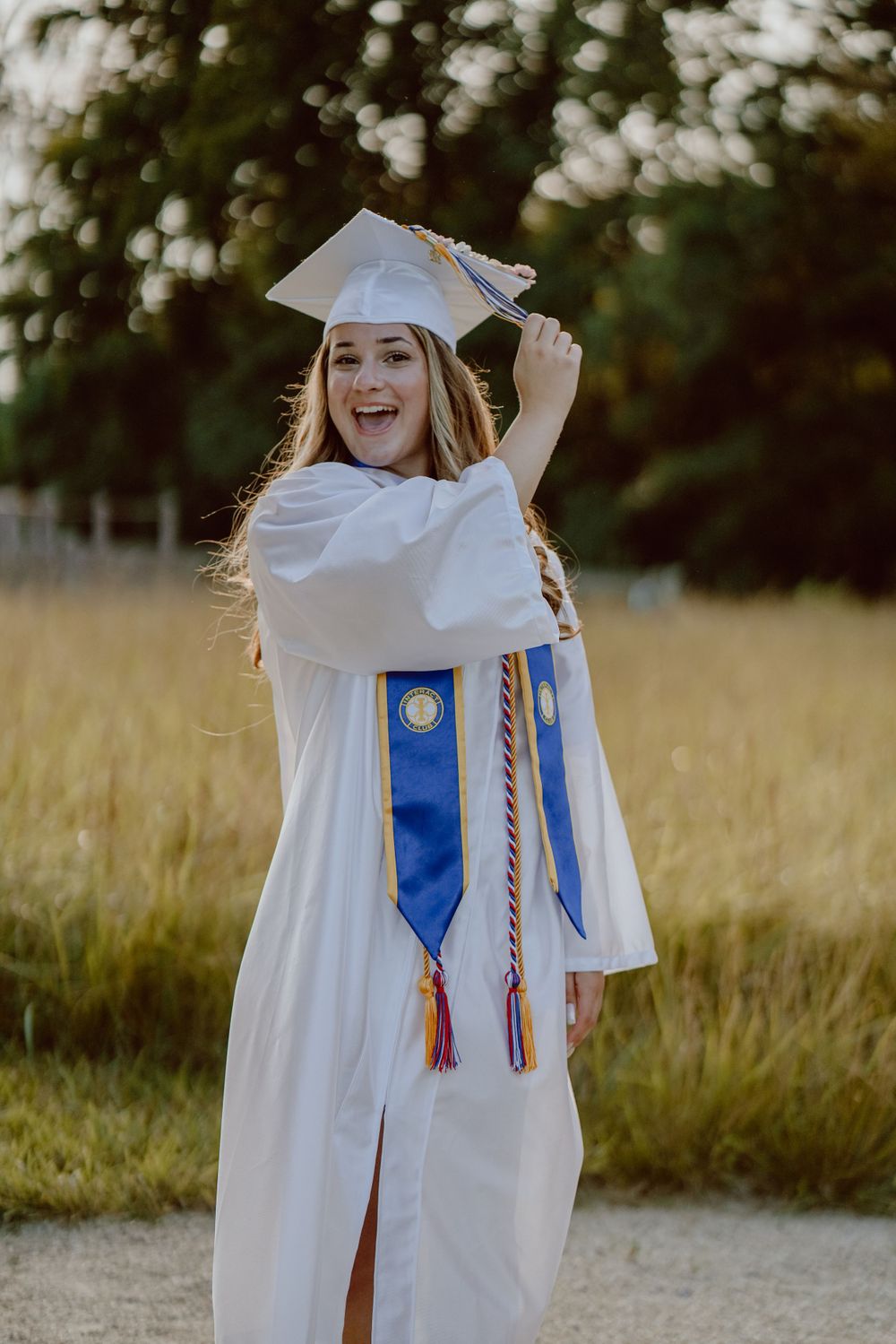 Graduate in white cap and gown with blue stole and honor cords walks through a sunlit meadow.
