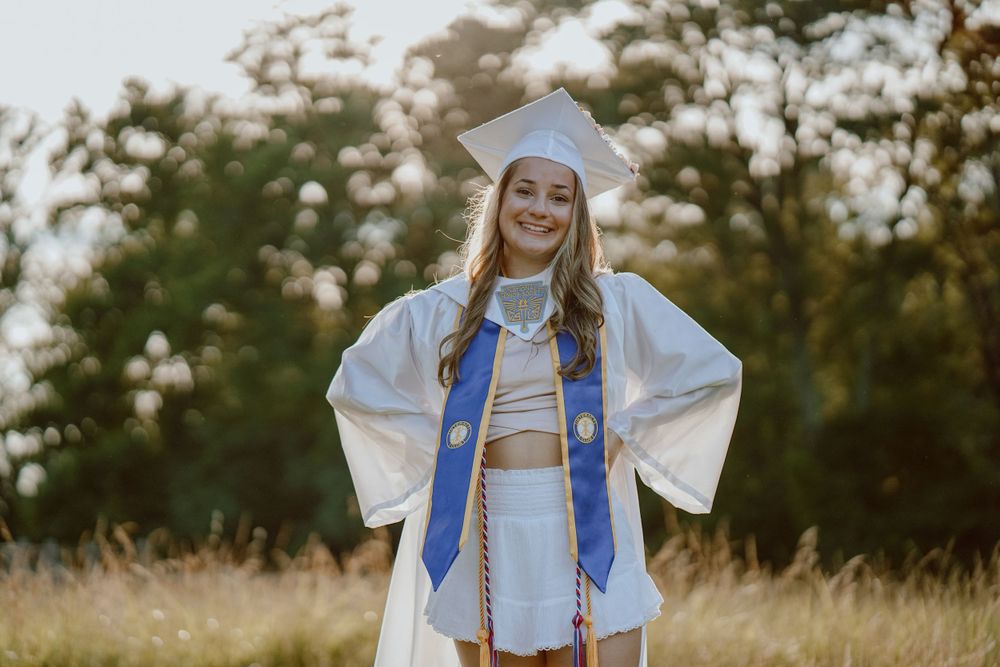 A graduate in white cap and gown with blue stole stands confidently in evening light.