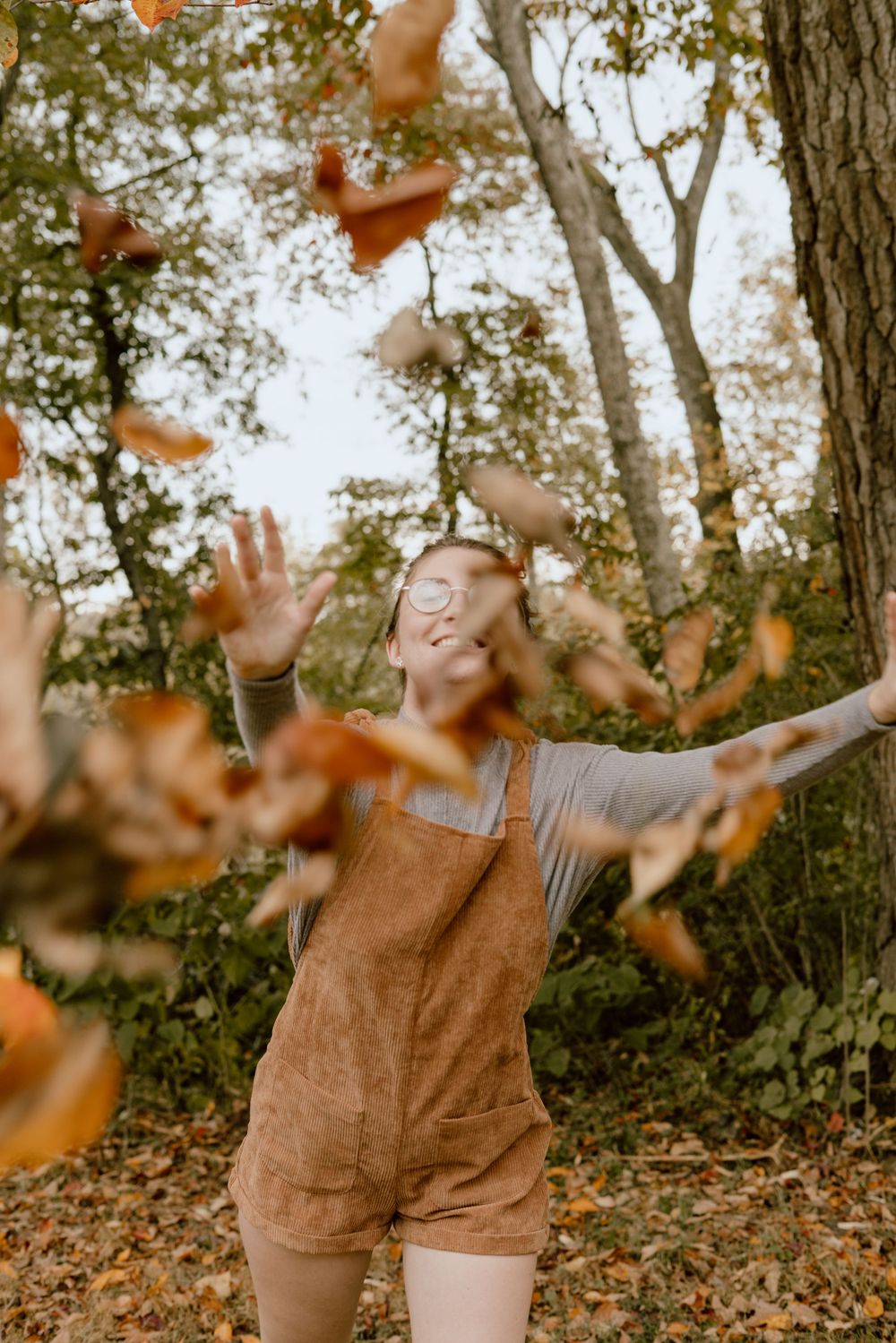 Person in brown overalls plays joyfully with autumn leaves in a wooded setting.