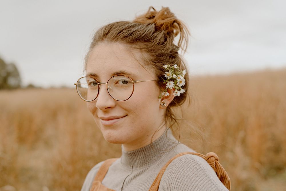 Portrait in a golden field wearing round glasses and small flowers in hair.