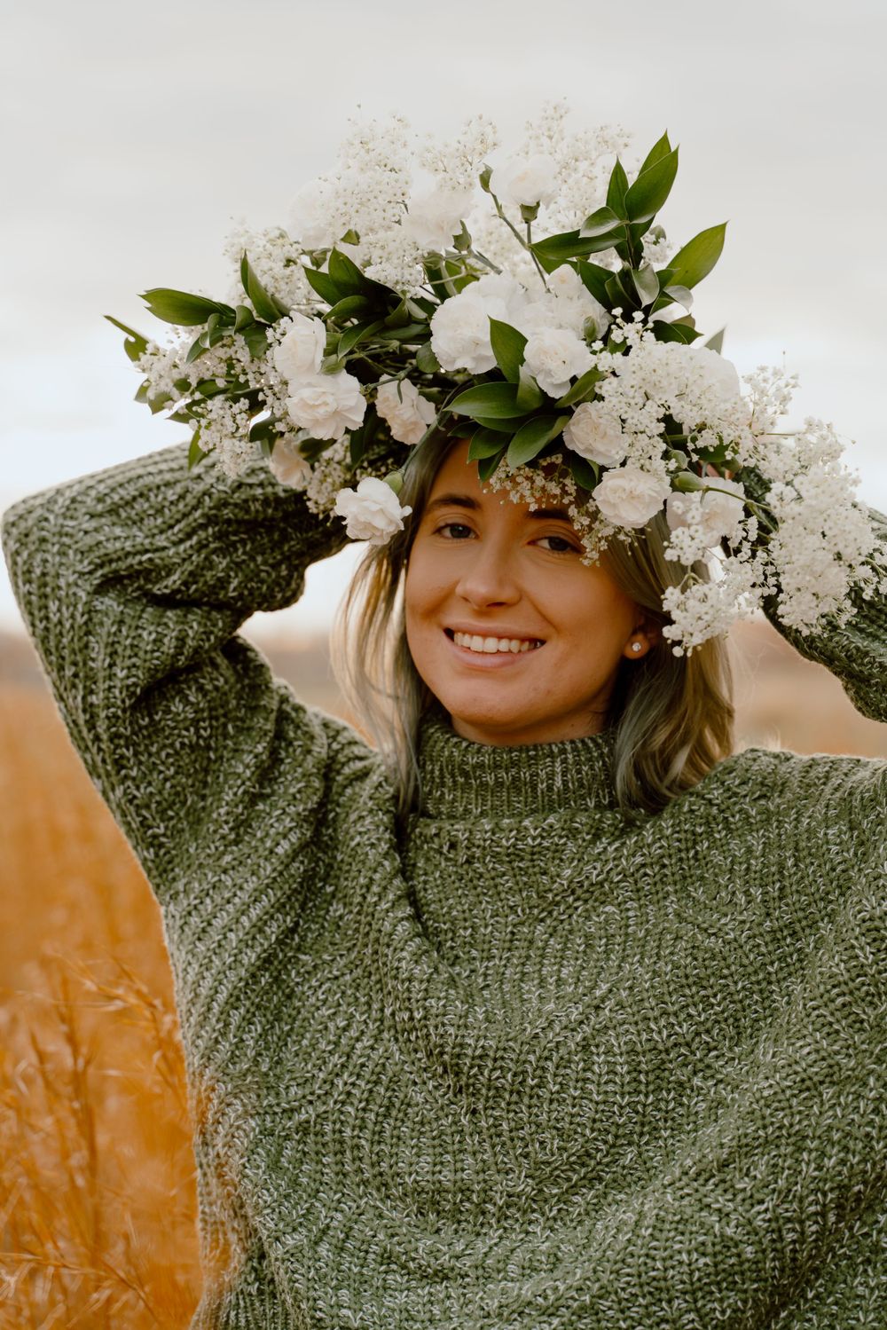 A delicate white floral crown adorns a cozy green knit sweater in an autumn field setting.