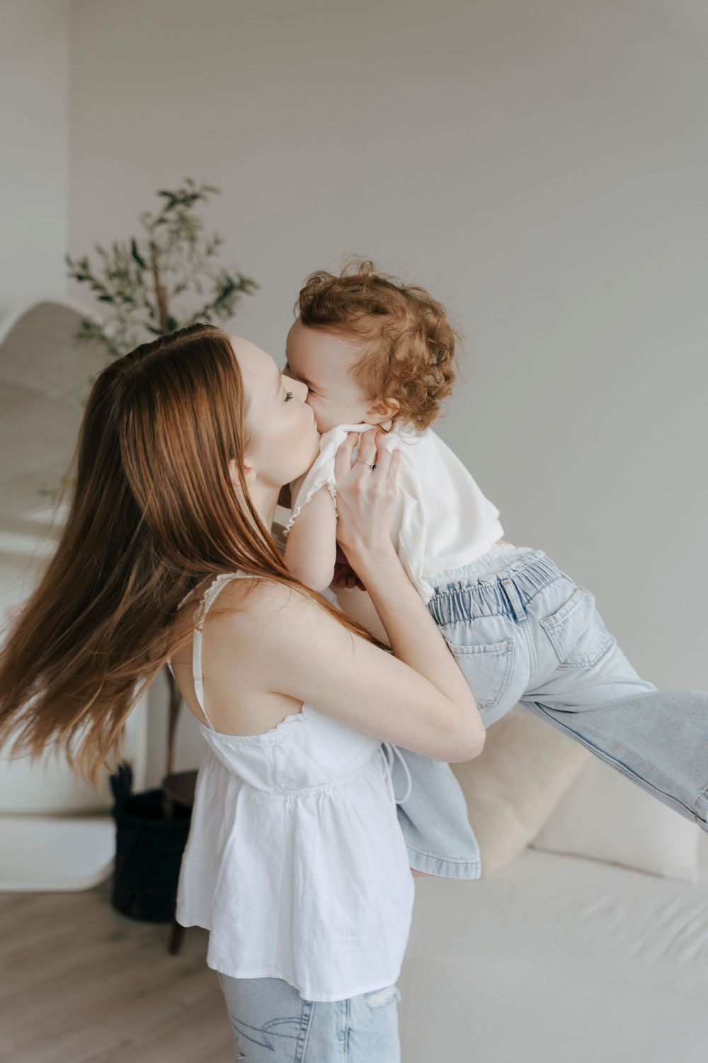A young mother lovingly kisses her curly-haired toddler while holding them up in a bright, modern room.