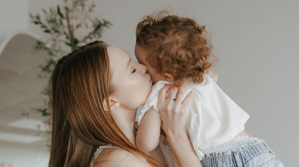 A loving mother gently kisses her curly-haired toddler in a bright, airy room.
