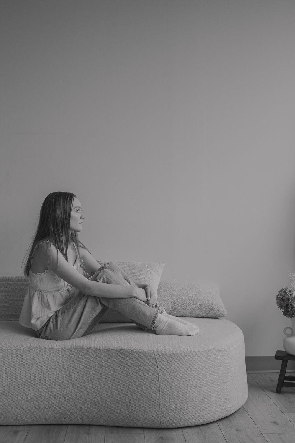 Black and white photo of a young woman sitting on a curved sofa, gazing thoughtfully to the side.
