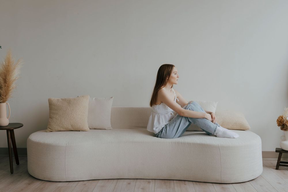 Young woman sitting cross-legged on a modern beige curved sofa in a minimalist living room with neutral decor.