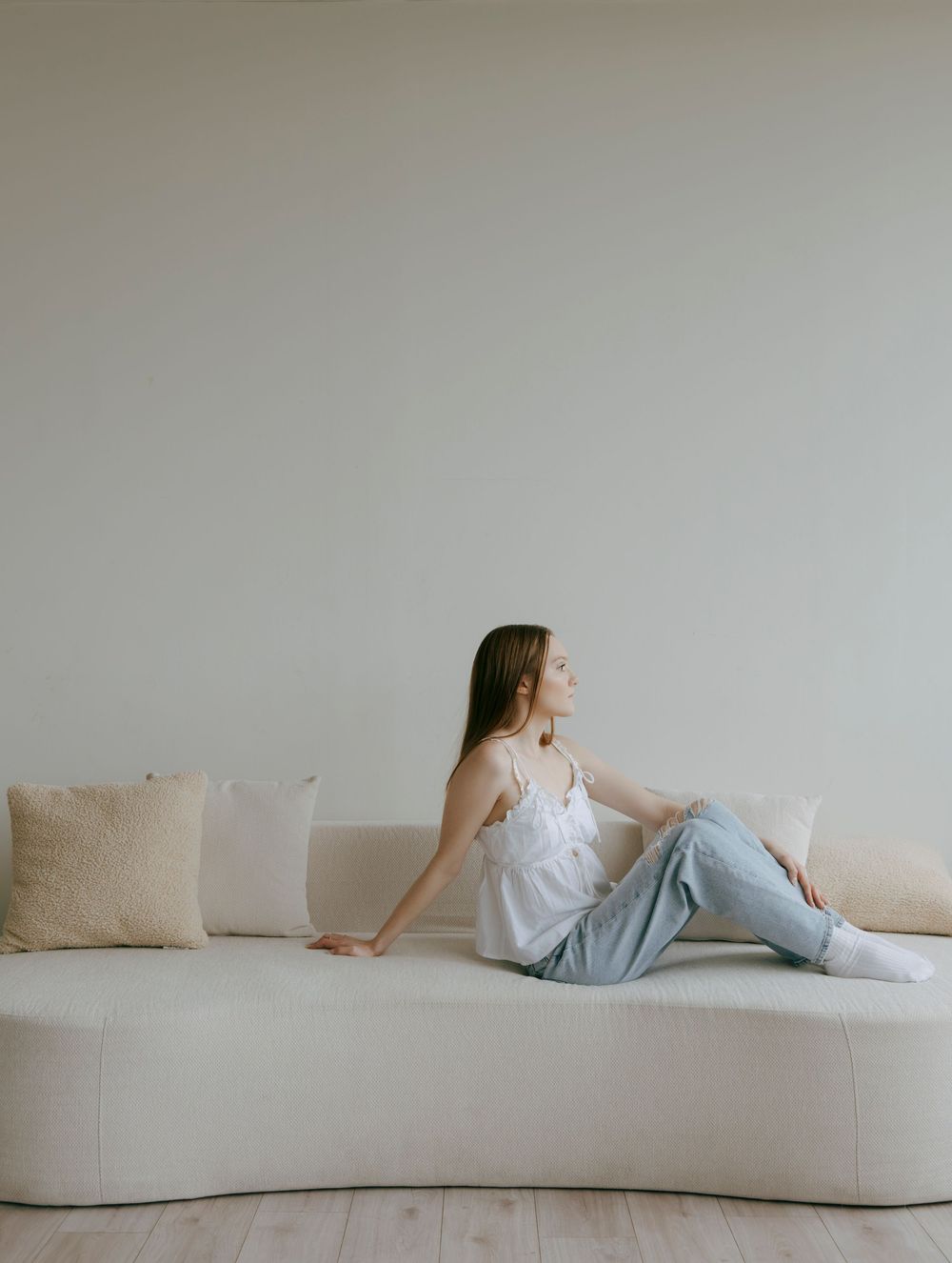 Young woman in white camisole and jeans relaxing on a curved cream sofa in a minimalist room.
