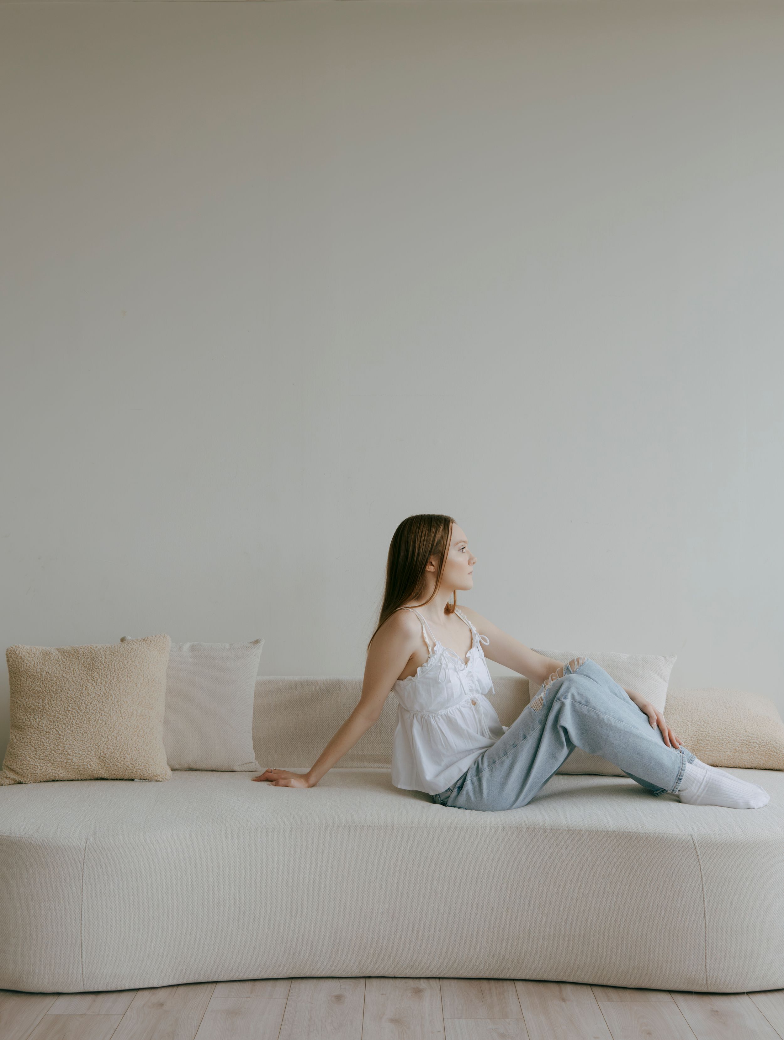 Young woman in white camisole and jeans relaxing on a curved cream sofa in a minimalist room.